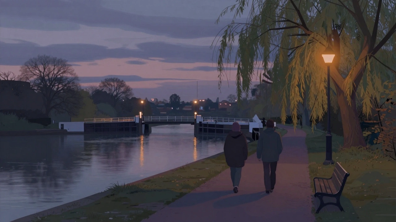 Silhouettes of two people walking along the Thames at dusk near Hounslow Lock, with distant airport lights reflecting on water.